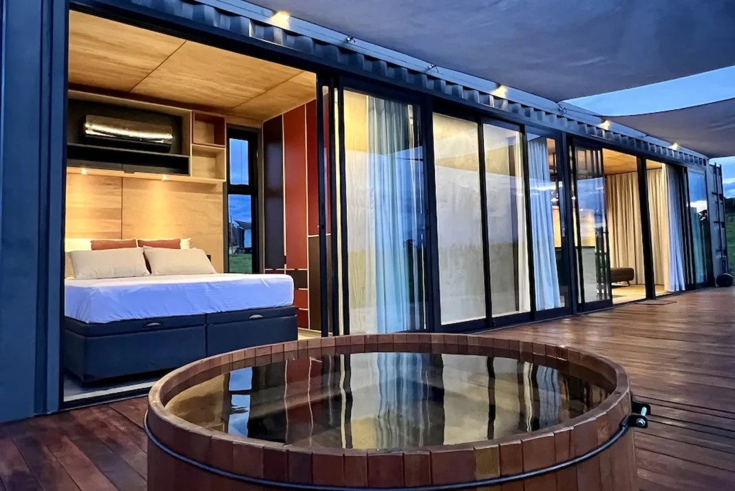 Moriá Village Uma banheira de hidromassagem de madeira redonda está em um deck de madeira. A banheira de hidromassagem está cheia de água e as portas de vidro deslizantes atrás dela estão abertas para mostrar um quarto com uma cama feita. Acima, um telhado branco com tiras pretas estende-se sobre o deck.